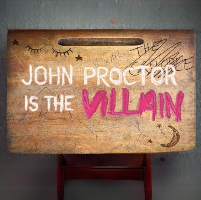 Image of a wooden desk with doodles on it and the text "John proctor is the Villain" written on it